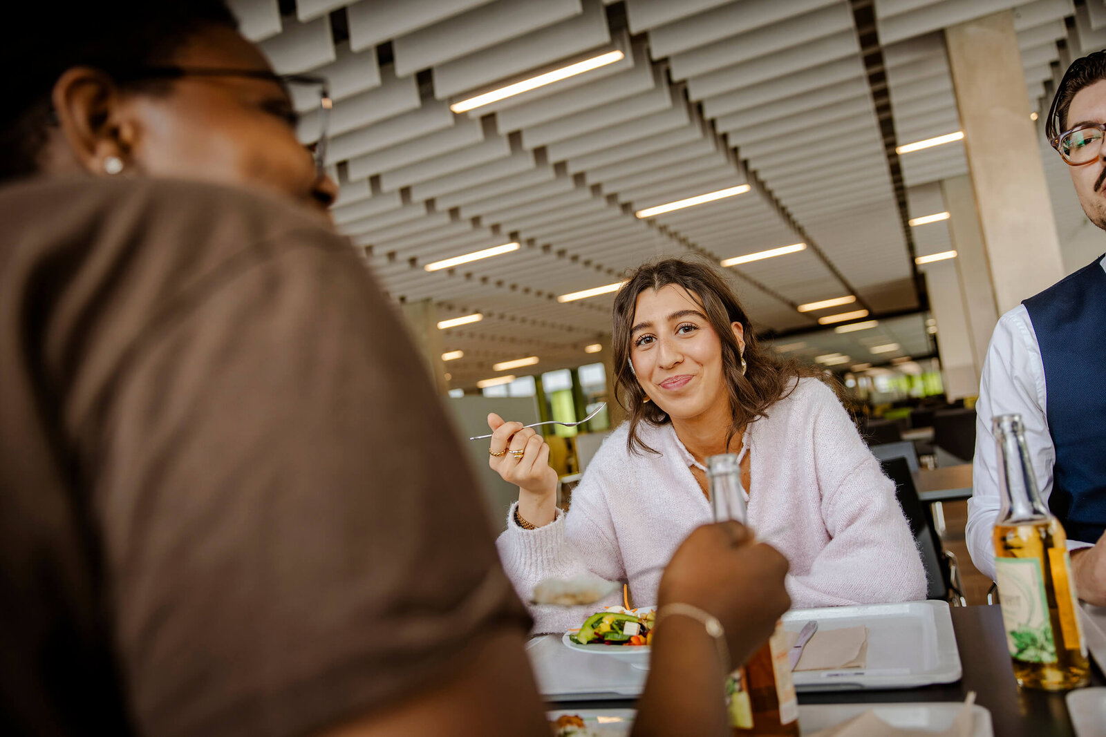 Drei Personen sitzen in einem Tisch in einer Mensa und essen, im Fokus ist dabei eine junge Frau, die eine Gabel in der Hand hält und lächelt.